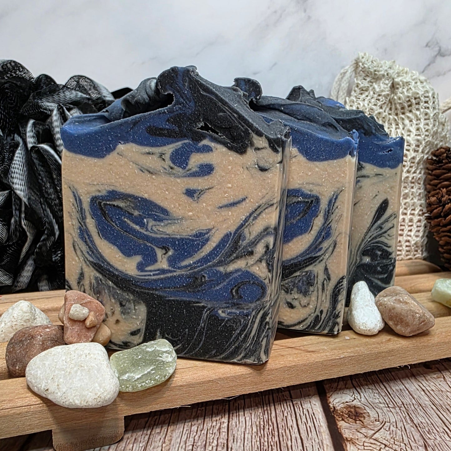 Three bars of soap with blue and beige swirls on a wooden surface with stones and a textured white background.