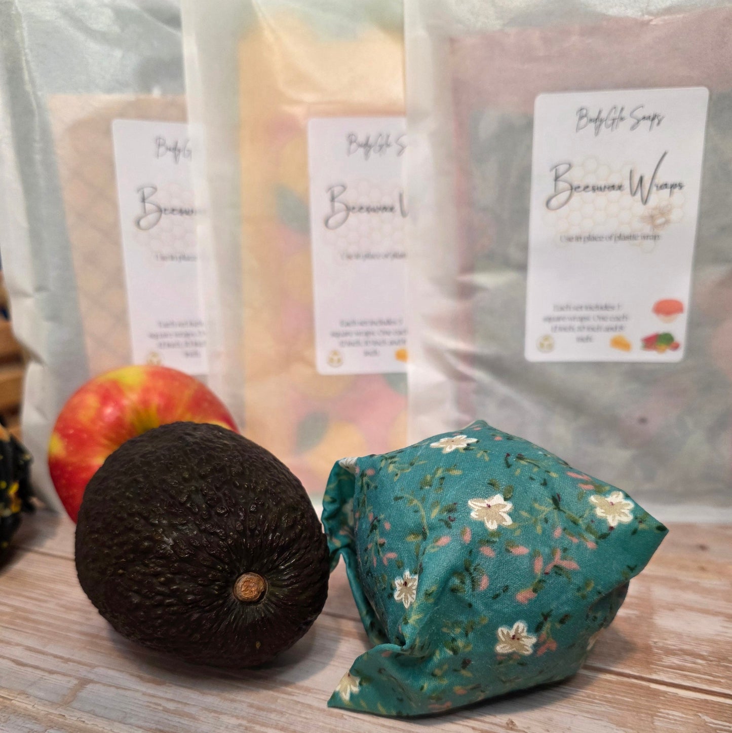 Reusable beeswax wrap with an avocado and apples, surrounded by wrapped produce bags on a wooden surface.