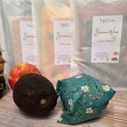 Reusable beeswax wrap with an avocado and apples, surrounded by wrapped produce bags on a wooden surface.