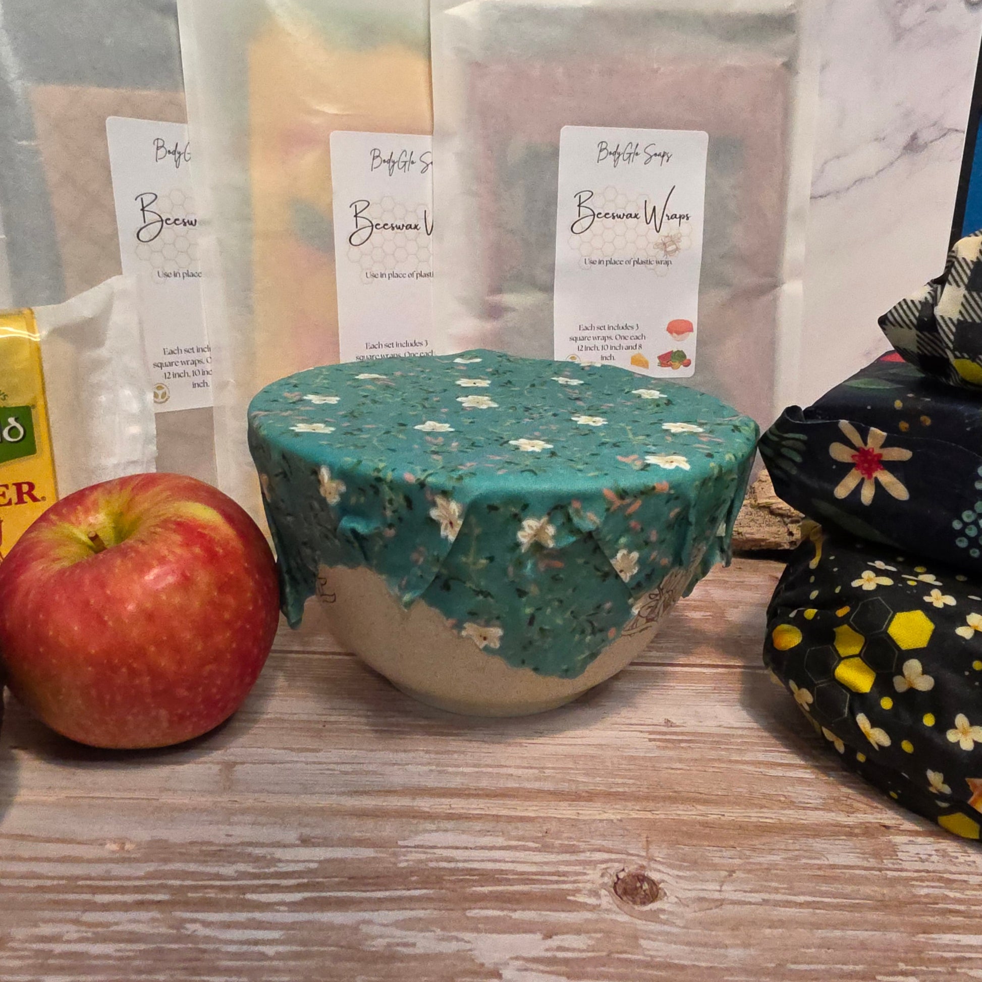 100% cotton beeswax wrapped bowl with floral pattern on a wooden surface, next to an apple and a package.