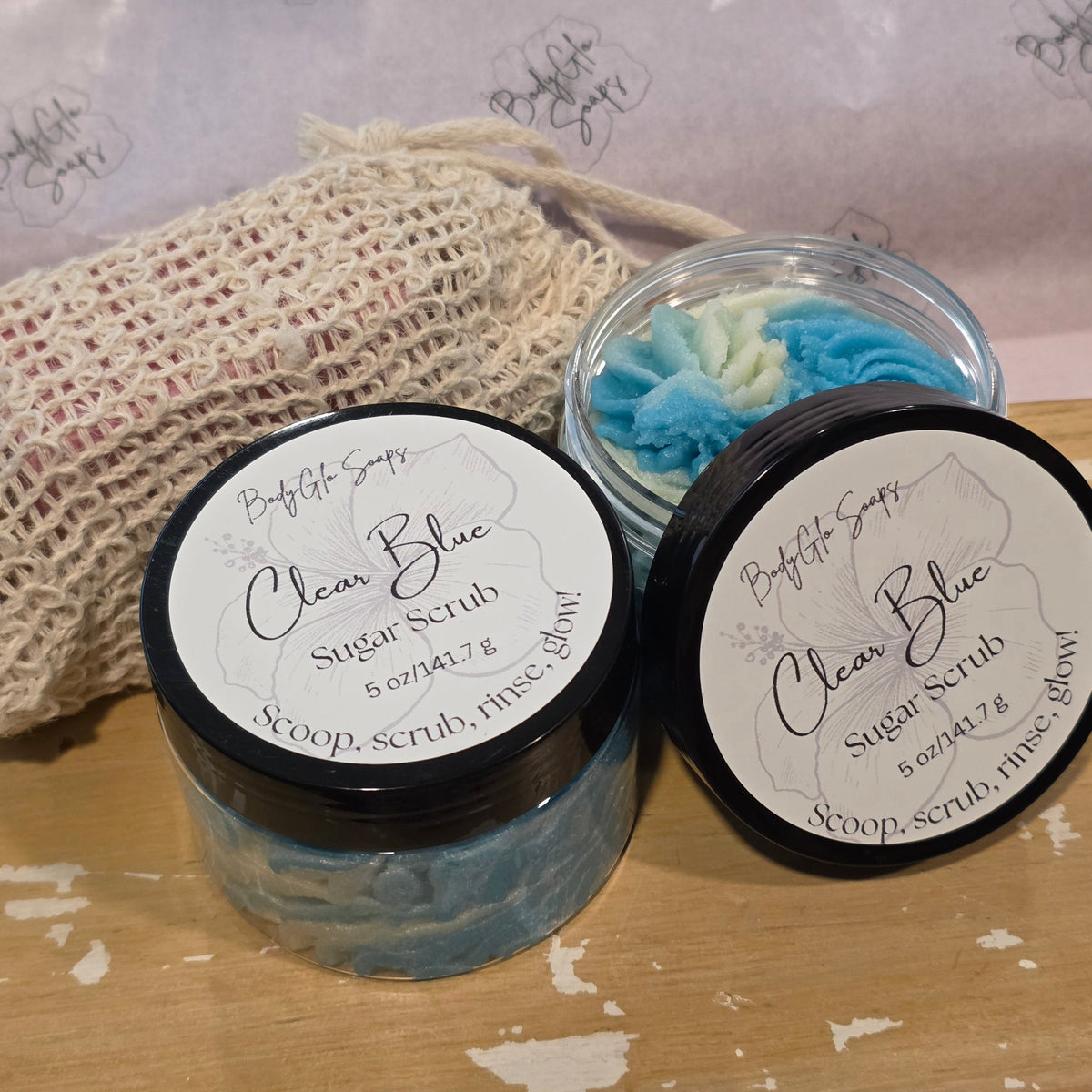Two jars of sugar scrub Clear Blue Sugar Scrub with black lids and white labels, one open jar showing blue and white scrub, and a beige mesh bag on a wooden surface.