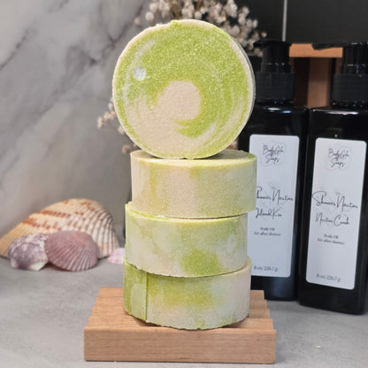 Four round bath salts stacked on a small wooden block, with one round standing upright on top. The bath salts are light green and cream marbled.