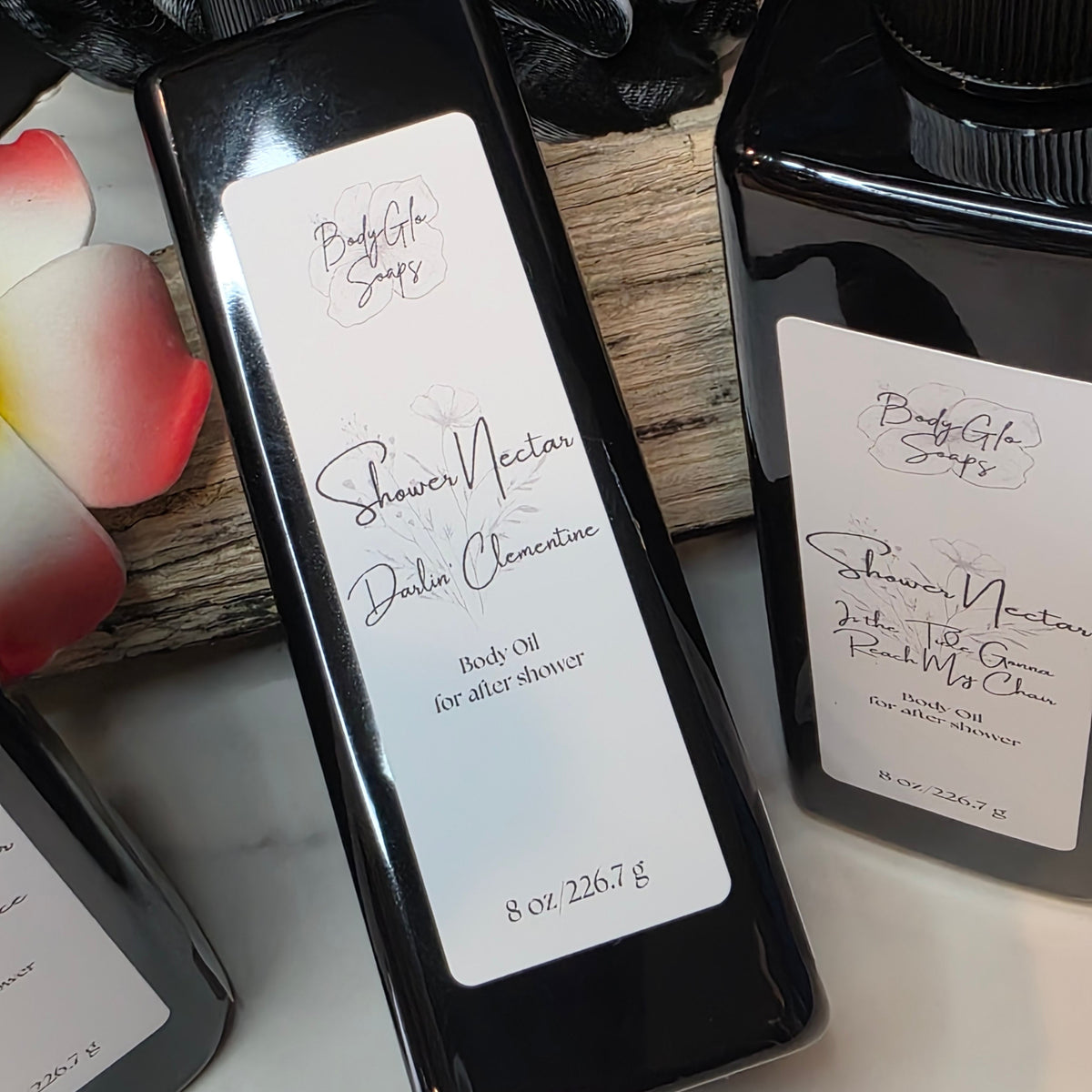 Three black bottles of shower products arranged on a white surface with a wooden board and black mesh in the background. The central bottle is upright and reads Shower Nectar Darlin' Clementine, with two other bottles partially visible on either side.