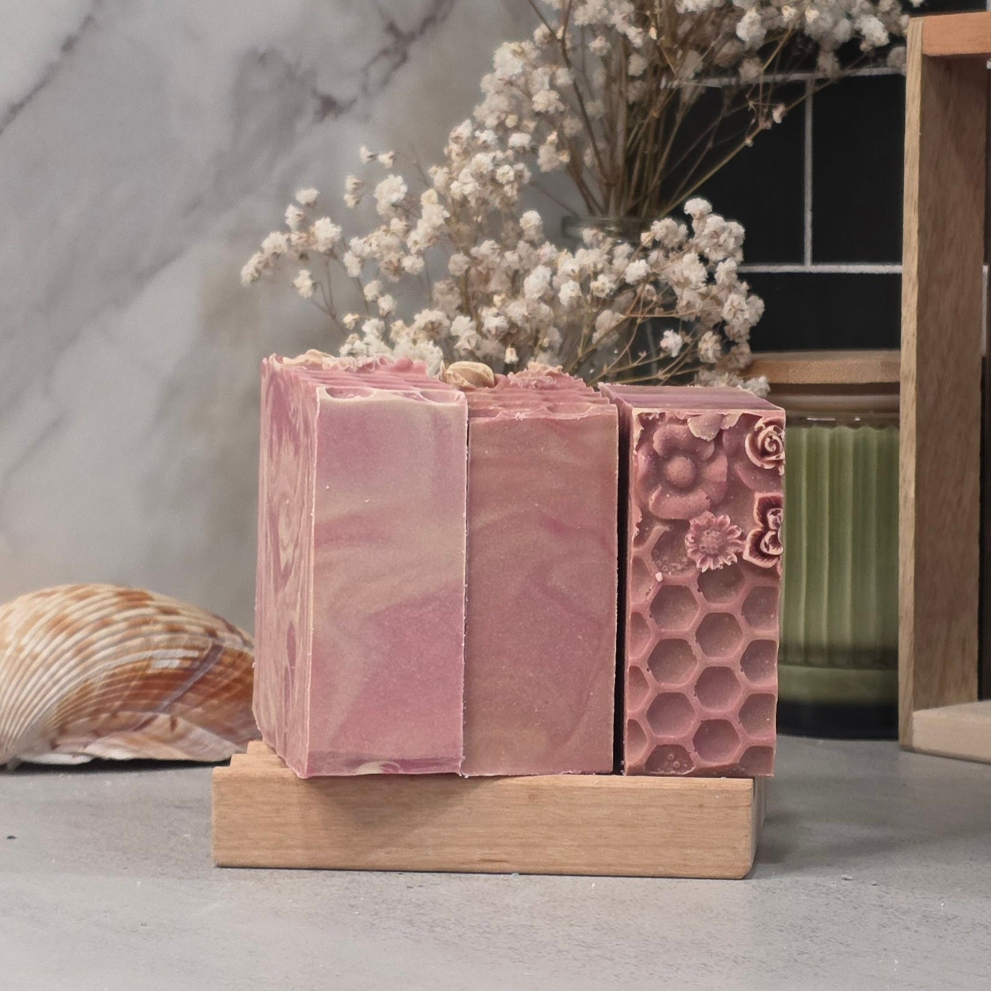 Three pink handmade soap bars displayed on a small wooden stand, with dried flowers, a seashell, and a marble-patterned background.