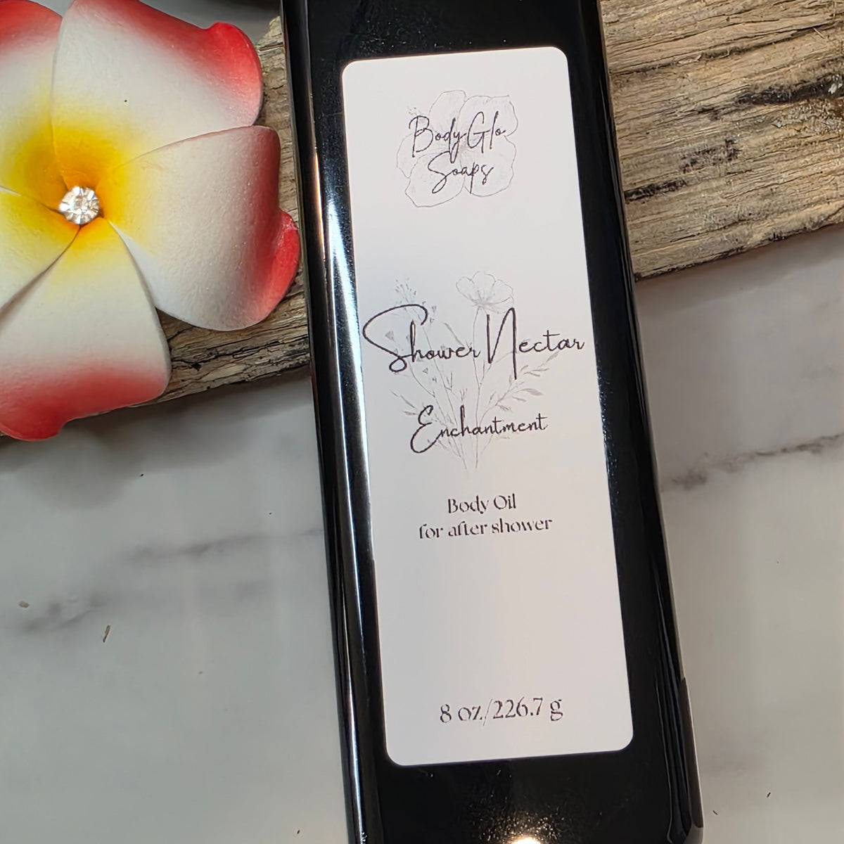 A black rectangular bottle of BodyGlo Soap Shower Nectar Enchantment body oil, featuring a white label, styled on a marble surface with a tropical flower and wood background.