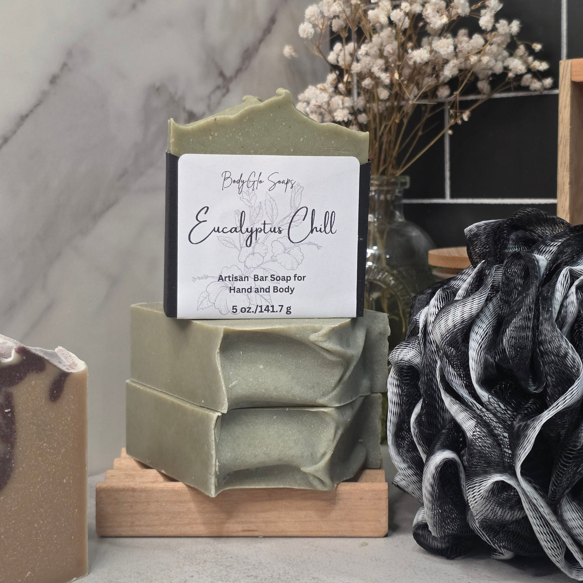 A handmade bar of soap labeled 'Eucalyptus Chill' by BodyGlo Soap, displayed on a wooden stand. The soap is a muted sage green color with a white paper label.