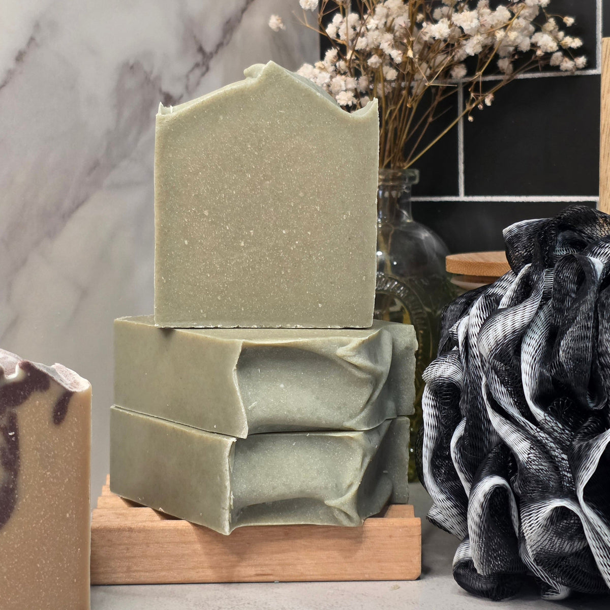 Three stacked handmade soap bars in a muted sage green color, displayed on a small wooden block with dried flowers and a black textured loofah in the background.