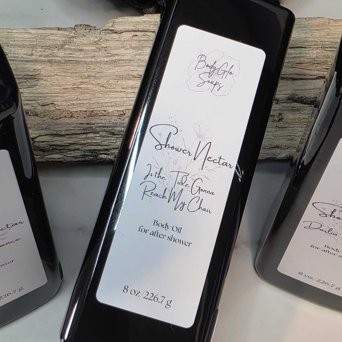 A bottle of Shower Nectar body oil for after shower, displayed on a white surface with rustic wood and other bottles in the background.