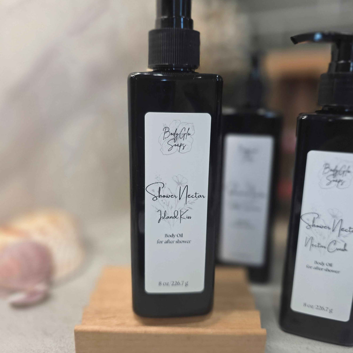 A black pump bottle labeled 'Shower Nectar standing on a small wooden block, with two matching bottles blurred in the background.