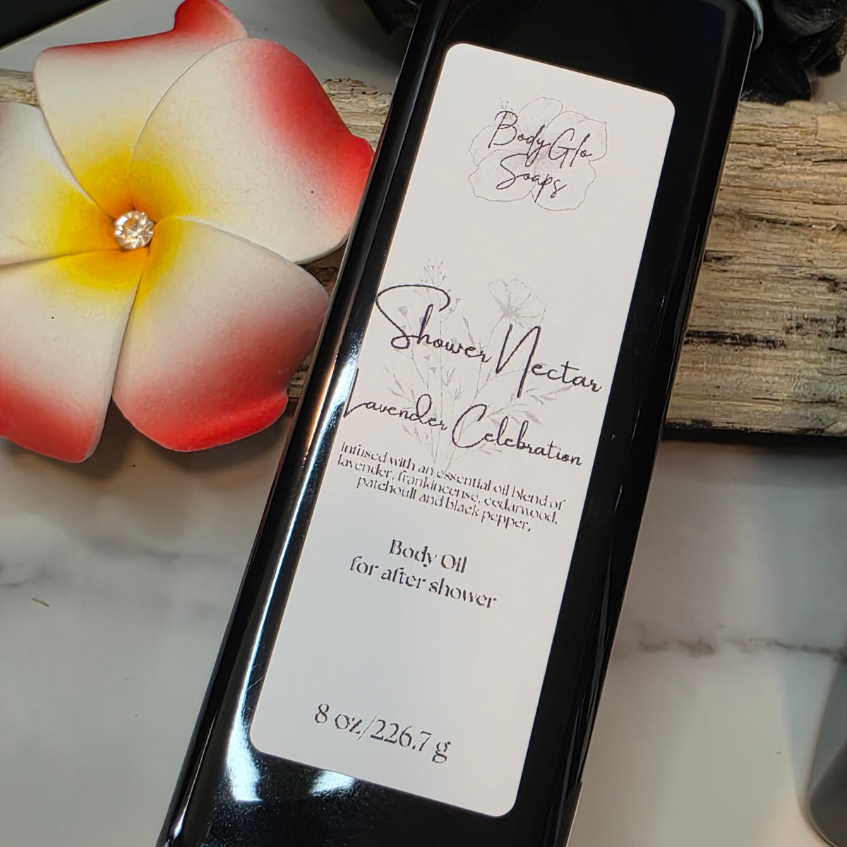 A black bottle of BodyGlo Soaps Shower Nectar Lavender Celebration  Essential Oil body oil, placed diagonally on a light marble surface next to a red and yellow plumeria flower.