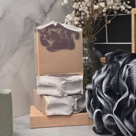 Three stacked handmade soap bars on a wooden stand, with dried white flowers in a glass vase and a black-and-white textured fabric prop in a styled bathroom-like setting.