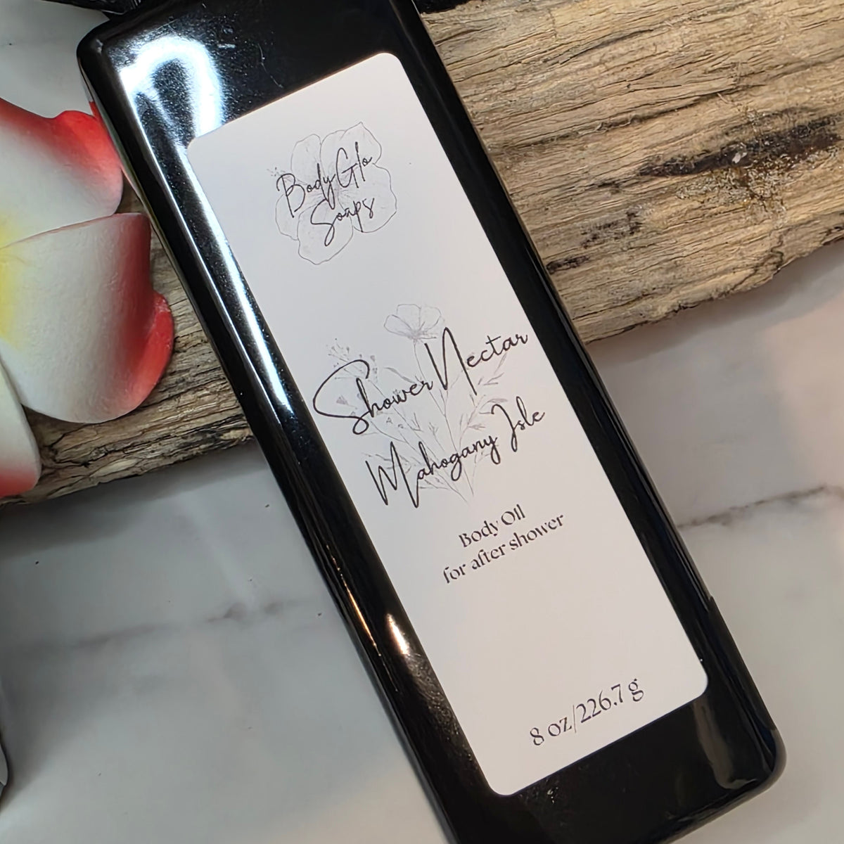 A black bottle of  Shower Nectar (Mahogany Isle) bath oil for after shower, resting on a piece of wood over a white marble surface.
