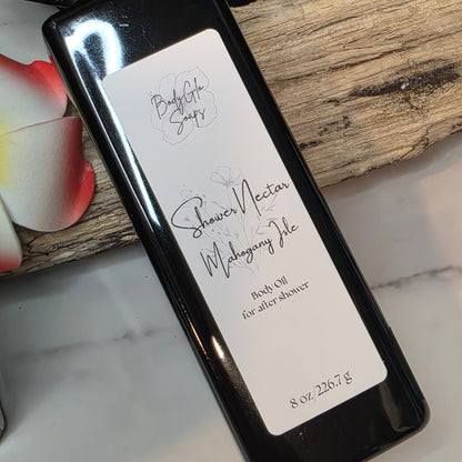 A black bottle of  Shower Nectar (Mahogany Isle) bath oil for after shower, resting on a piece of wood over a white marble surface.