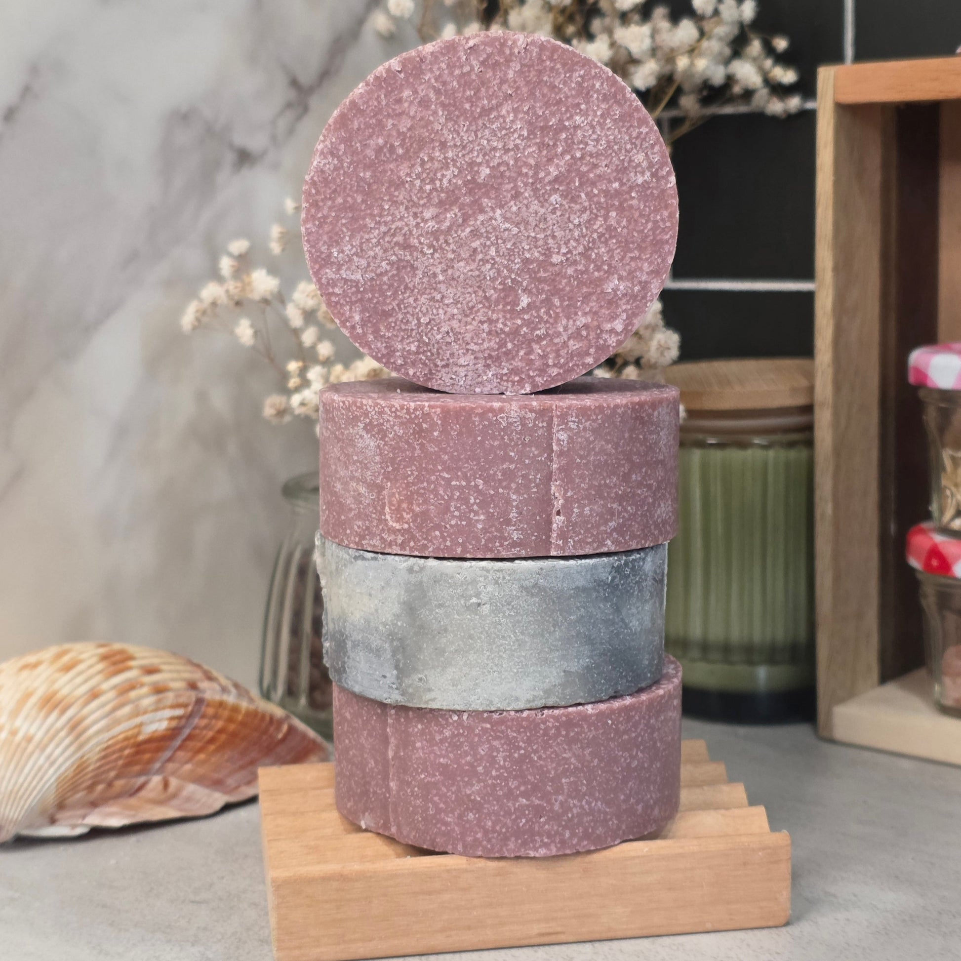 Three stacked rectangular bars of soap with a circular top piece, displayed on a small wooden stand. The soap bars have a dusty mauve-pink color with fine glitter, and the middle bar has a metallic silver finish.