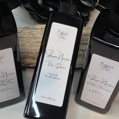A black plastic bottle Shower Nectar Pear Dance , flanked by two more bottles on a white surface with a rustic wood prop and black fabric in the background.