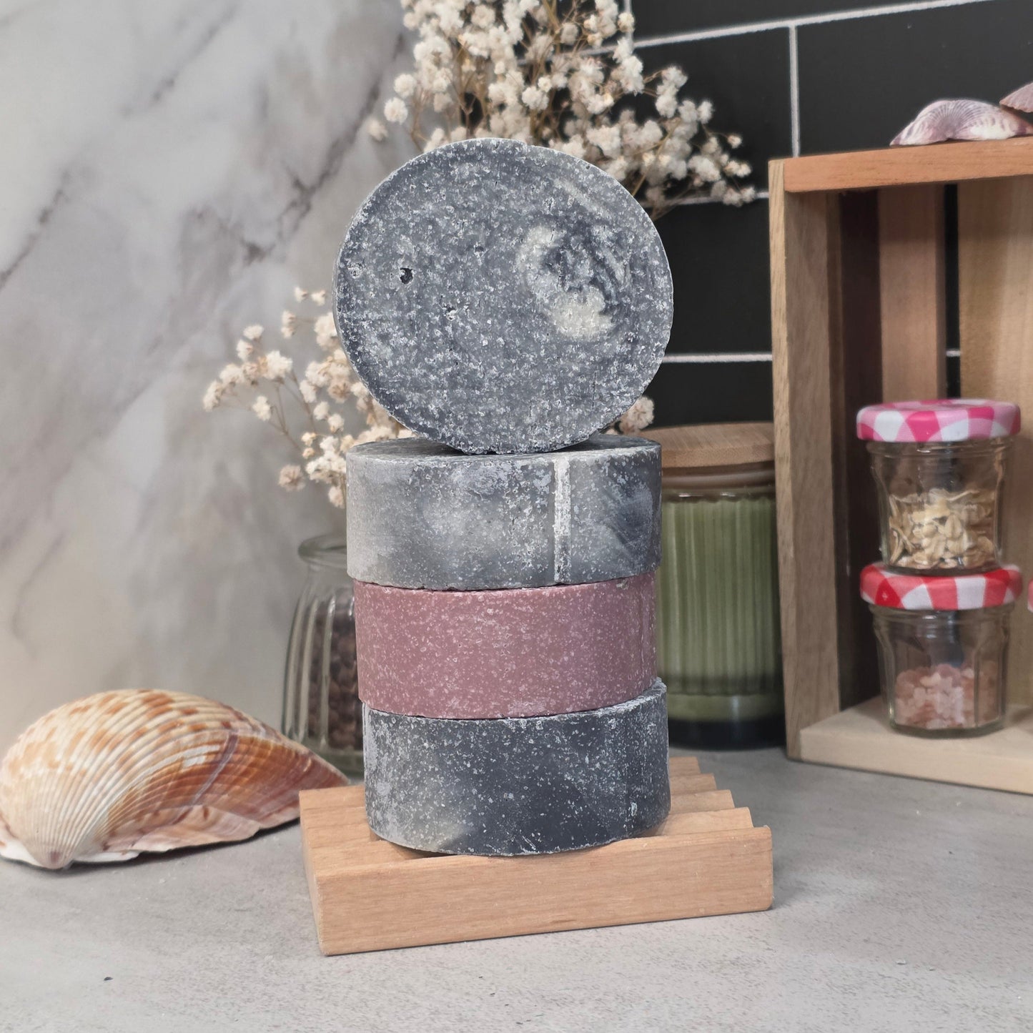 Three stacked rectangular bath salts or soap bars with a circular lid resting on top, displayed on a small wooden stand. The top piece is dark gray with white speckles, the middle piece is dusty rose pink, and the bottom piece is dark gray with white speckles.