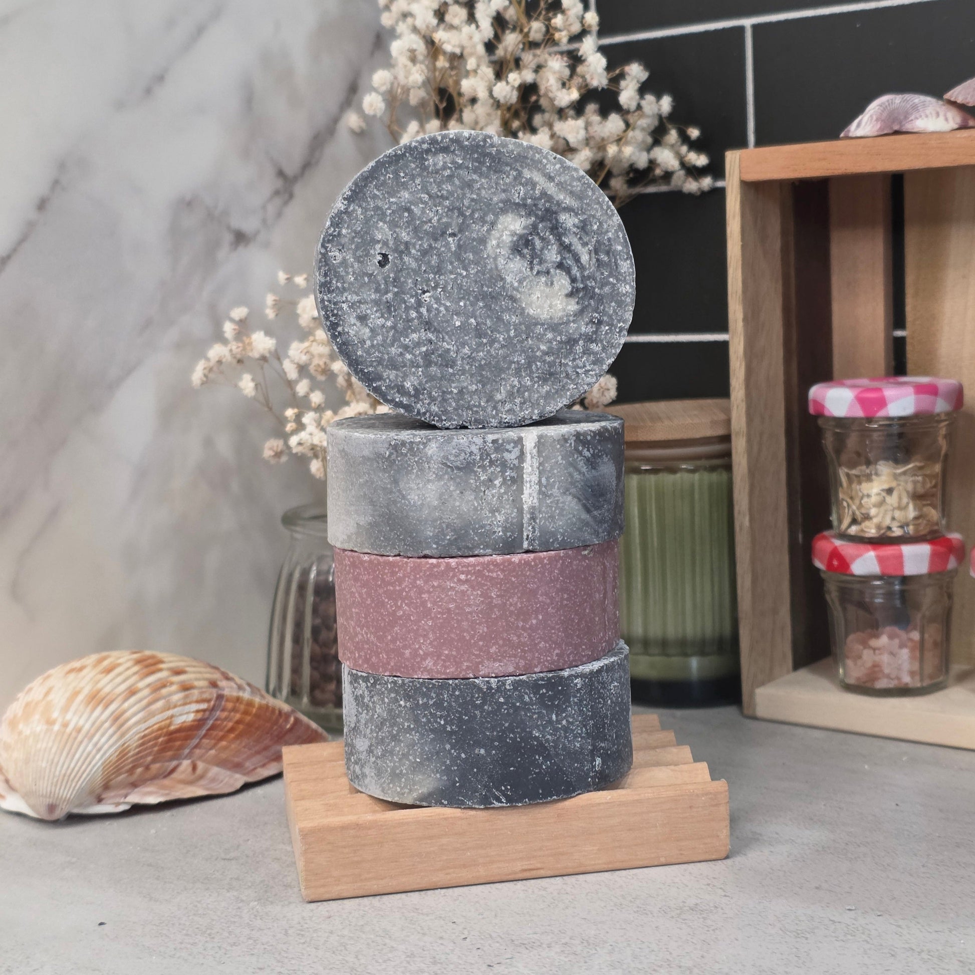 Three stacked rectangular bath salts or soap bars with a circular lid resting on top, displayed on a small wooden stand. The top piece is dark gray with white speckles, the middle piece is dusty rose pink, and the bottom piece is dark gray with white speckles.