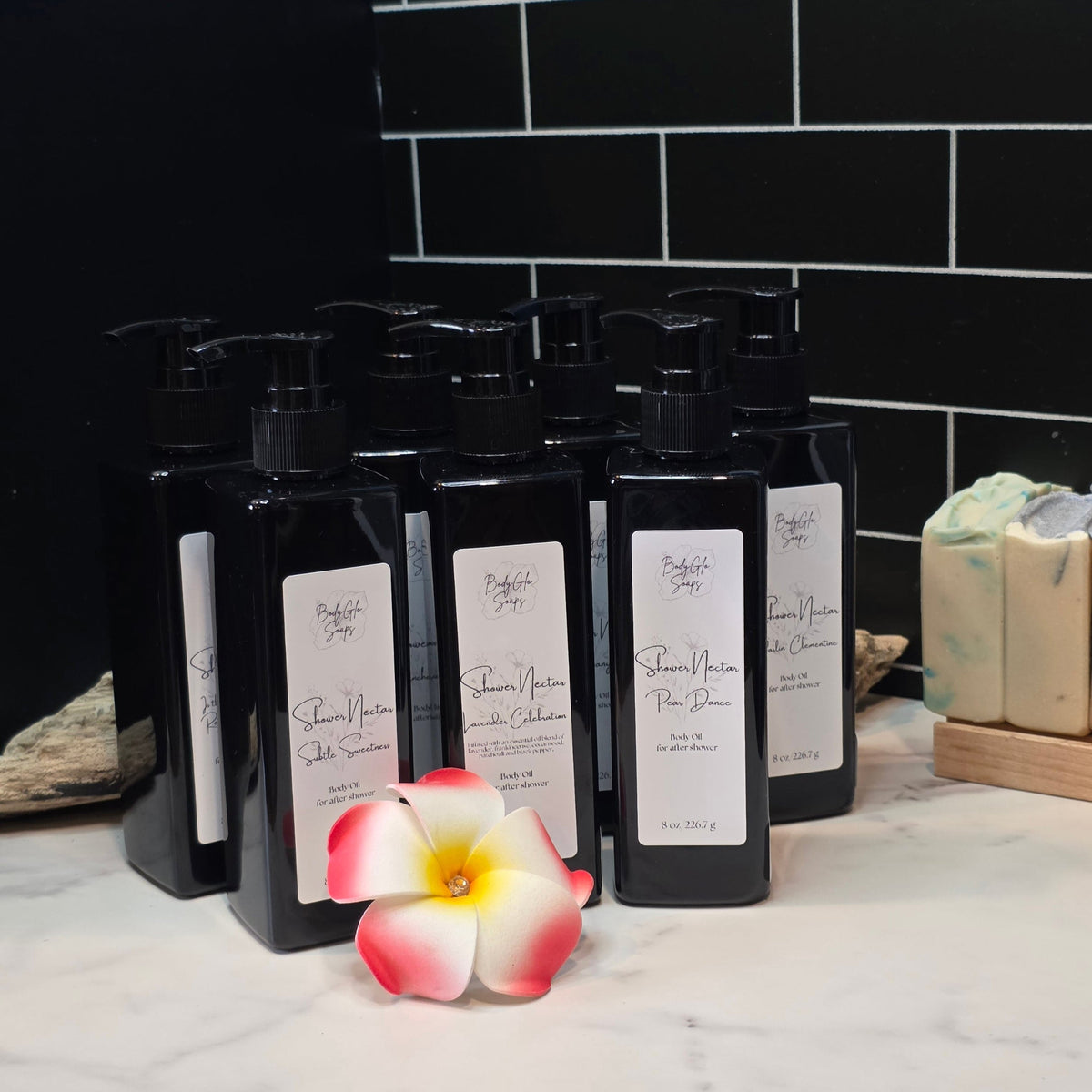 A set of black pump bottles with white labels from the brand 'BodyGlo Soaps' arranged on a white marble surface against a black subway tile wall. A pink and white plumeria flower is placed in front of the bottles, and a bar of soap sits on a wooden stand to the right.