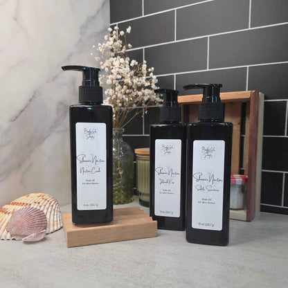 Three black pump bottles of shower oil arranged on wooden blocks on a countertop, with a marble wall, dark subway tile, dried flowers, and decorative shells in the background.