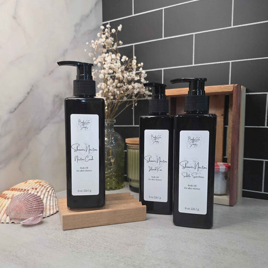 Three black pump bottles of shower oil arranged on wooden blocks on a countertop, with a marble wall, dark subway tile, dried flowers, and decorative shells in the background.