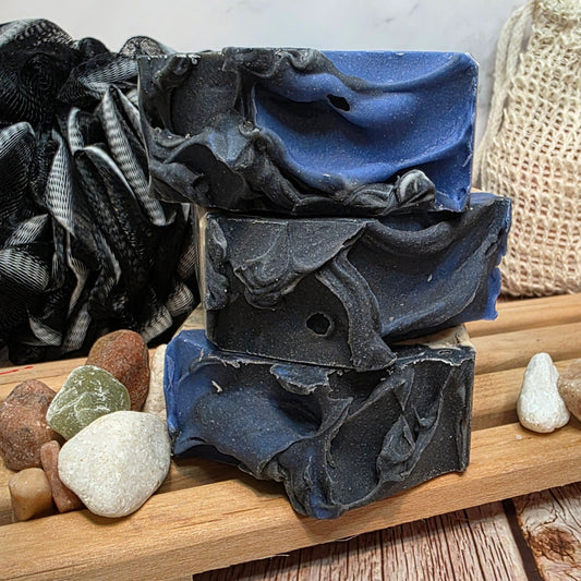 Stack of blue and black soap bars on a wooden tray with pebbles.