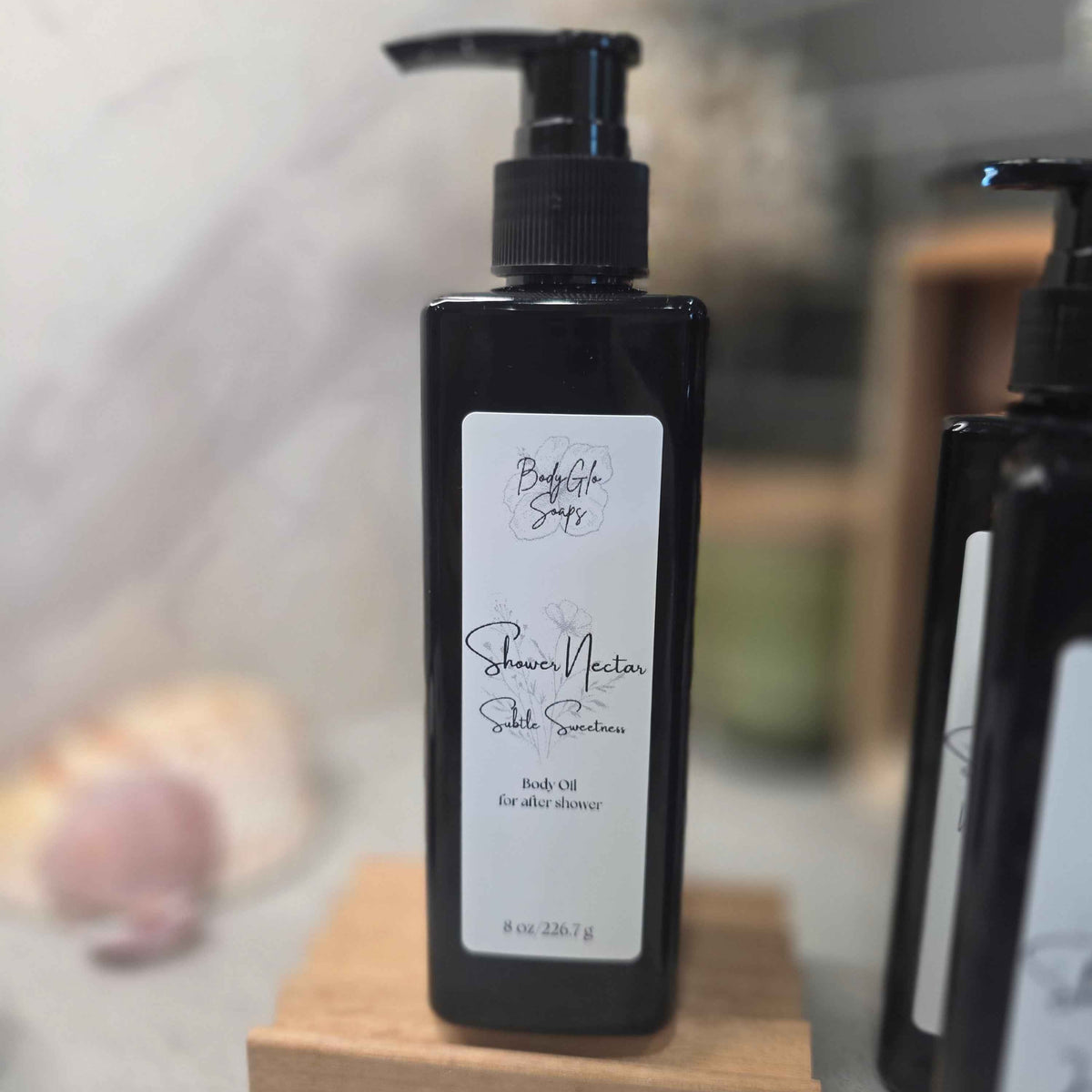 A black pump bottle of Shower Nectar Body Oil for after shower use, standing on a small wooden block. The label is white with black text and logo.