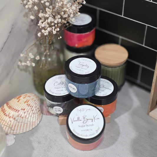 A group of five scented sugar scrub jars in various colors, arranged on a countertop next to a glass vase with dried baby's breath and a seashell. The background features a black subway tile wall.