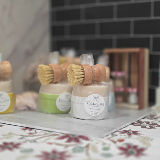 Dish soap bars in ramekin with wooden-handled brushes, displayed on a marble tray over a floral surface with a dark tiled wall in the background.