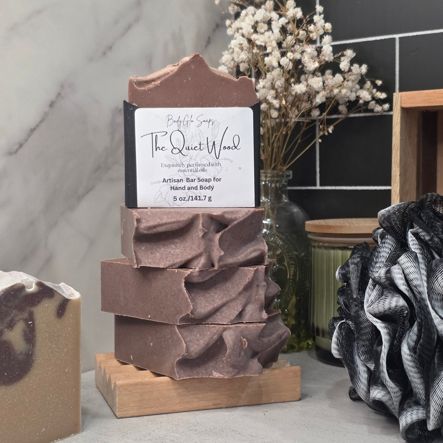 A stack of handmade soap bars in a rustic wooden holder on a countertop, with a front bar labeled 'The Quiet Wood' . The setting includes dried flowers, a dark tiled wall, and other bath/spa items.