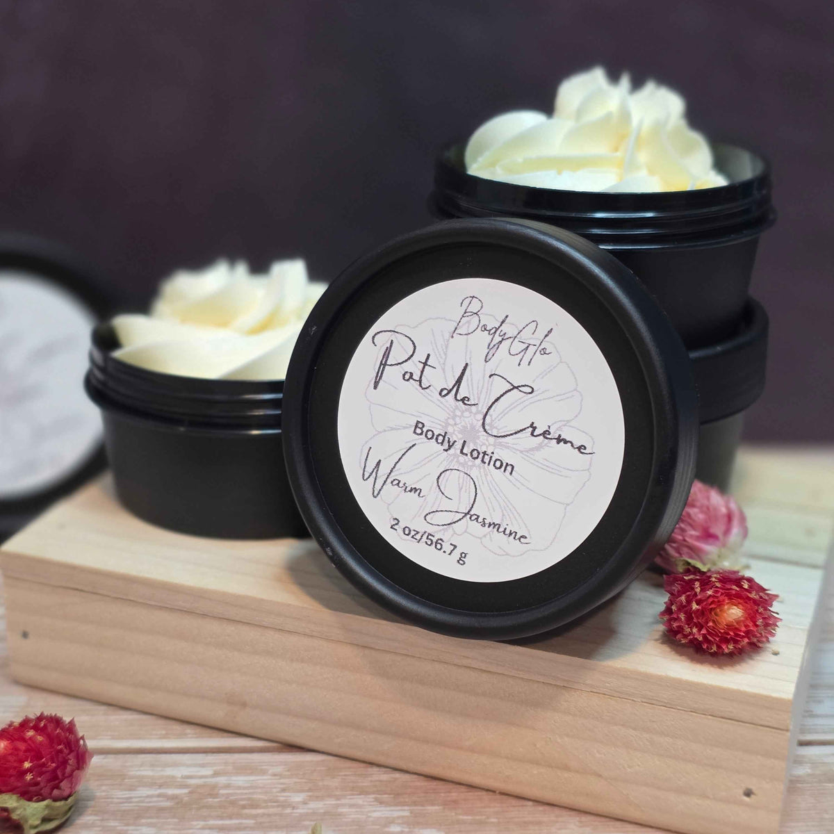 A black round jar of body lotion with a white circular label reading  Pot de Creme' and 'Jasmine', styled on a wooden board with dried flowers against a dark background.