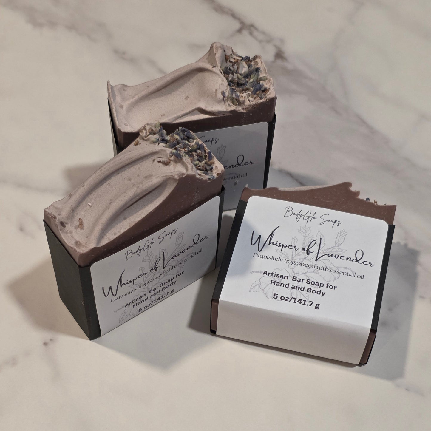 Three artisanal bar soaps from BodyGlo Soaps, including 'Whisper of Lavender', displayed on a light marble surface. One soap is shown uncapped to reveal its textured, speckled lavender-infused top, while two are in their dark cardboard packaging with white labels.