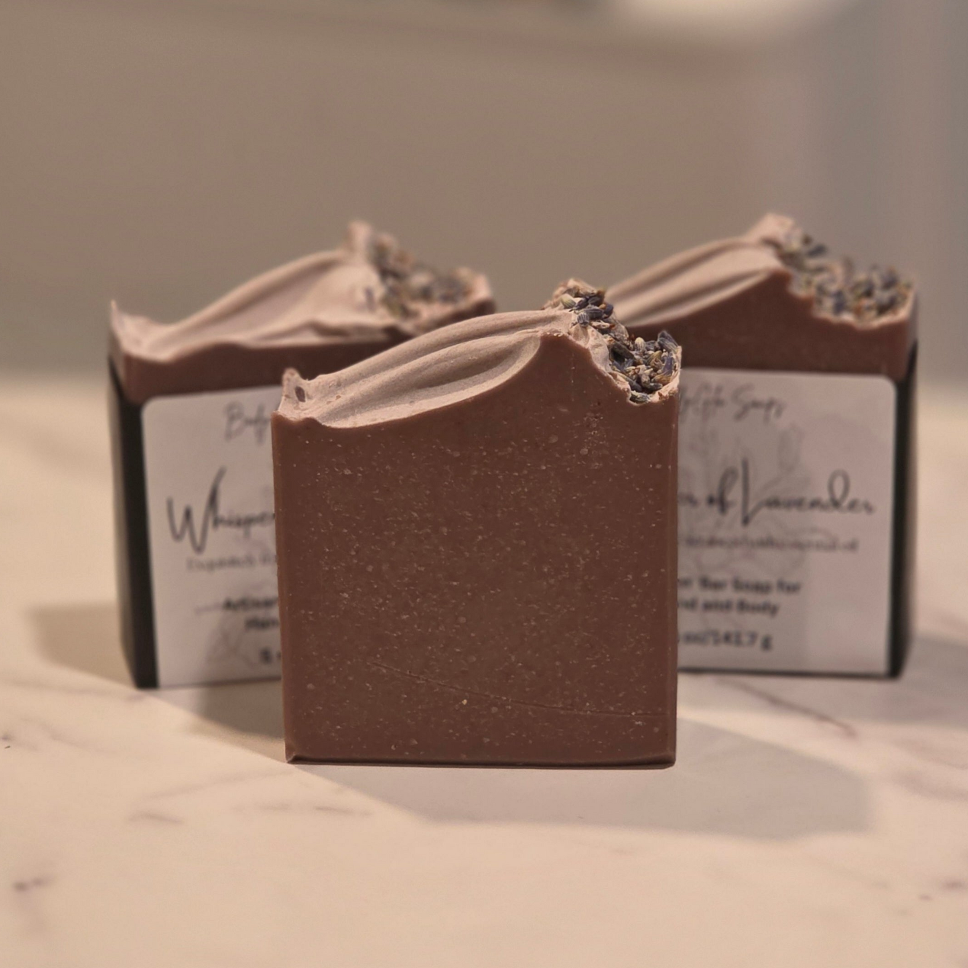 Three bars of handmade lavender soap displayed on a light marble surface, with one bar in the foreground and two in the background inside their black and white packaging.