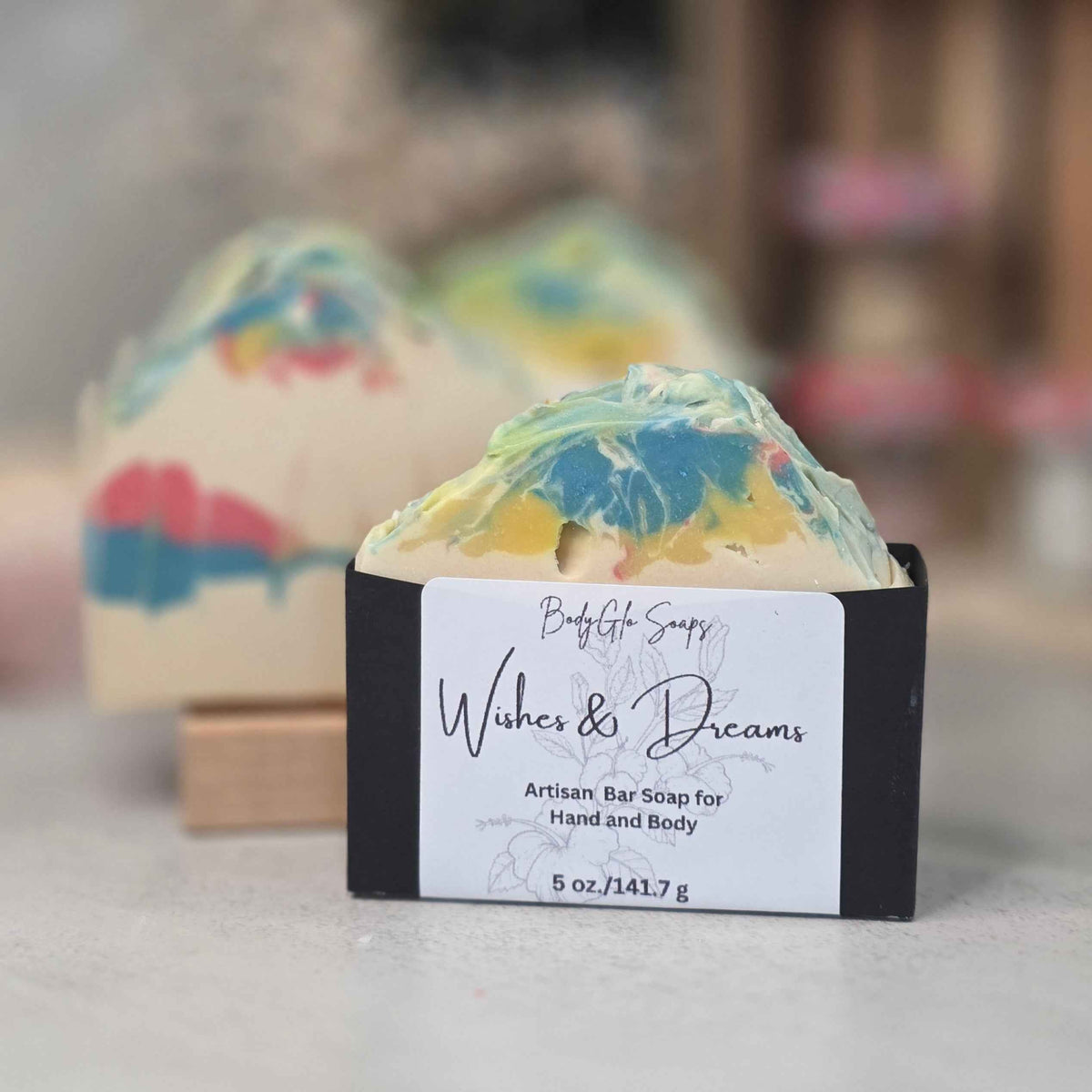 A handcrafted artisan bar soap bar wrapped in a black paper sleeve with a white label reading 'Wishes & Dreams'  placed in front of two more soap bars on a light gray surface with a blurred background.