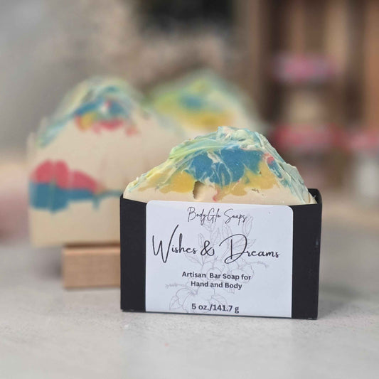 A handcrafted artisan bar soap bar wrapped in a black paper sleeve with a white label reading 'Wishes & Dreams'  placed in front of two more soap bars on a light gray surface with a blurred background.
