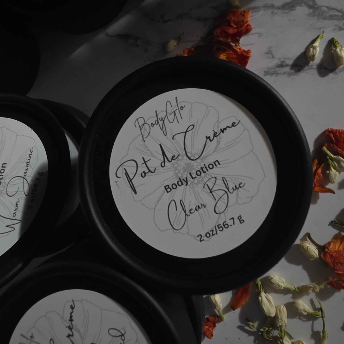 Black round cosmetic jars with white circular labels arranged on a light marble-like surface with dried orange and white flowers around them. The labels show handwritten text including 'Body Lotion'